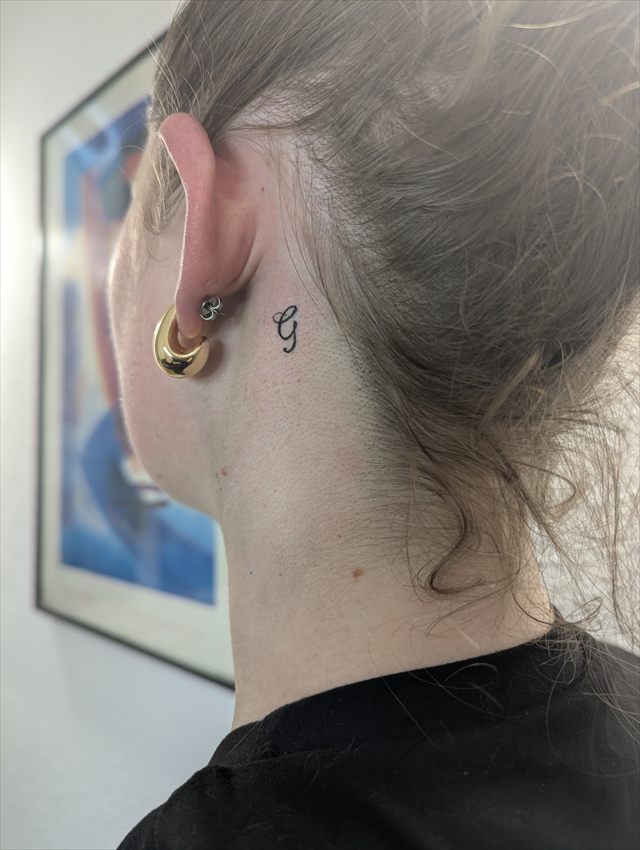 Tiny script letter tattoo behind an ear